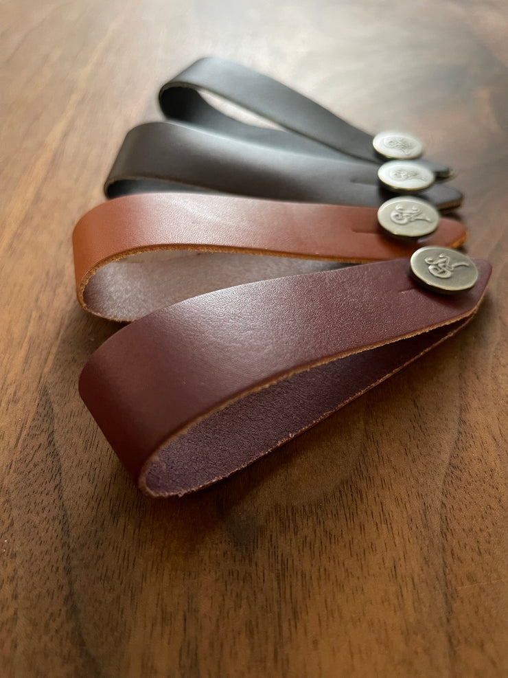 Leather guitar headstock adapter straps in black, brown, and dark brown with metal logo buttons